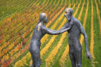 Two human figures as sculptures stand in front of colorful vines in an autumnal atmosphere,