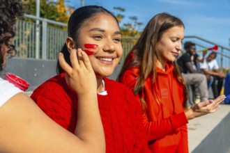 Young happy spectators sitting in bleachers having face paint applied before a sports event,