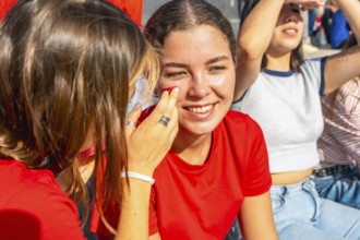 Young women at a sunny stadium, one friend painting red face paint on anothers cheek as they smile