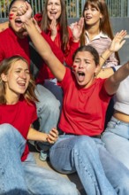 Young women sitting together, wearing red shirts and face paint, passionately cheering and