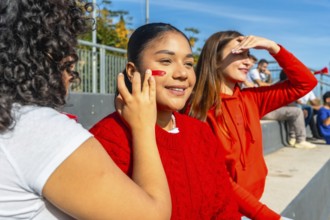 Young happy female friends enjoying a game at an outdoor stadium, painting faces and cheering for