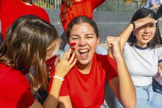 Young female friends in red team colors and face paint are thrillingly watching a sports event,