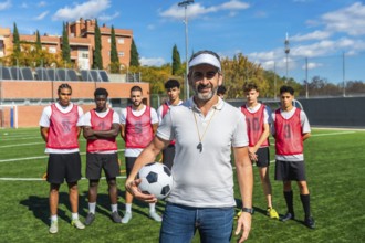 Male coach with a confident smile standing on a football field, holding a soccer ball, with his