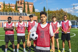 Diverse group of male soccer players in red bibs standing on a training pitch, holding a ball and