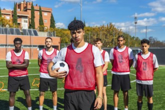 Multi ethnic group of young men in red bibs standing on a green football pitch, preparing for