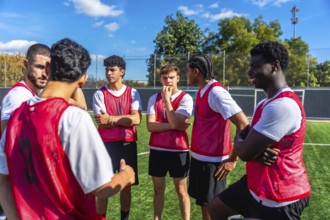 Young male soccer players wearing red bibs huddling on a green artificial turf field, listening