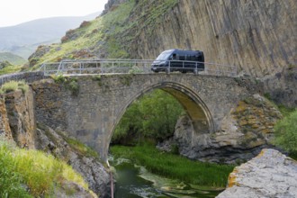 A black van drives over an old stone bridge, surrounded by picturesque rocky and river landscape,