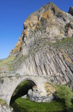 A high stone bridge stretches in front of an impressive rock formation under a bright blue sky,