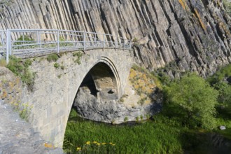 View of a stone bridge across a clear river with lush greenery and dramatic rock formations in the