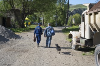 Two men walking a dog on a rural road flanked by old vehicles and green surroundings, police
