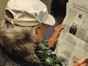 Elderly woman reading in the newspaper with a sticker on her cap from the cross-party citizens'