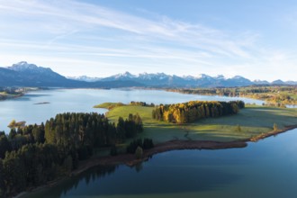Wide alpine landscape with lake and wooded hills in the morning light, surrounded by snow-capped