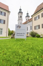 Sign for the Center for Psychiatry with two church towers in the background on a green field, ZFP