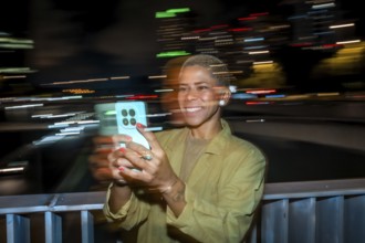 Woman smiling and holding a smartphone, capturing the vibrant motion blur of city lights at night,