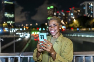 Woman smiling while interacting with her mobile phone, standing on a bridge with blurred city