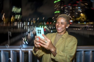 Smiling woman with short blond hair takes a nighttime smartphone selfie in an urban setting, bokeh