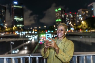 Happy woman smiling, holding a smartphone and enjoying a vibrant urban night view from a bridge,