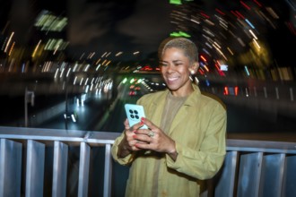 Smiling young adult woman using a mobile phone at night on a city bridge, enjoying digital