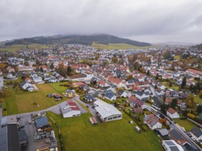 View of a city with colorful roofs surrounded by green fields and wooded hills under gray clouds,