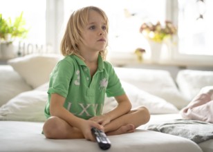 Child, boy, 7 years old, blonde, sitting on sofa, with remote control in hand, watching TV,