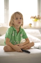 Child, boy, 7 years old, blonde, sitting on sofa, with remote control in hand, watching TV,