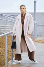 A woman stands confidently on outdoor steps, wearing a long light coat over a brown outfit. She