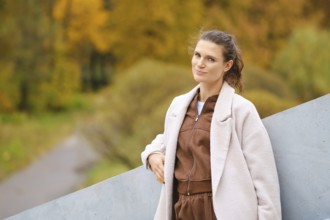 A woman with long hair stands confidently against a gray geometric structure. She wears a brown