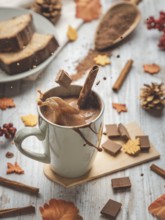 Hot chocolate splashes out of a cup surrounded by cinnamon sticks, cakes and autumn decorations