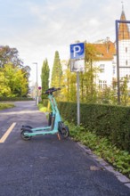 An electric scooter is parked on a sidewalk next to a parking sign against an autumn backdrop,