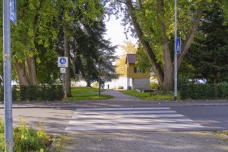 A pedestrian crossing leads to a park with tall trees and an autumn backdrop, Überlingen, Lake