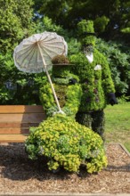 Live plant sculpture of Victorian man standing and woman with sun shade umbrella sitting on wooden