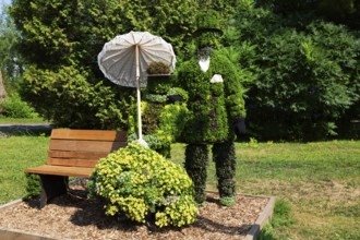 Live plant sculpture of Victorian man standing and woman with sun shade umbrella sitting on wooden