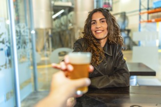 Woman smiling and raising a glass of frothy craft beer in a modern brewery, toasting an unseen