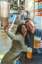 Happy young couple posing for a selfie with a smartphone, holding glasses of craft beer, and