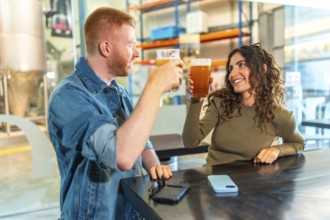 Happy young couple toasting craft beers and laughing together inside a modern brewery tasting room,