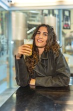Smiling woman with curly hair wearing a leather jacket, happily holding a glass of pale beer while