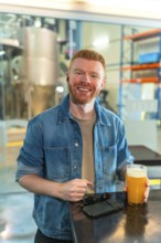 Smiling man enjoying a glass of craft beer in a modern industrial brewery. He stands wearing a
