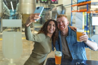 Young couple smiling for a selfie with craft beers in hand at a modern brewery tasting room,