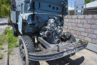 An old lorry with an open bonnet and no front body parts stands close to a brick wall in the sun,