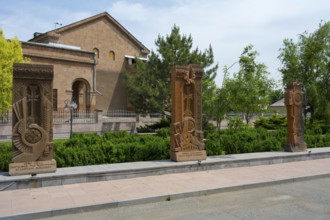 Three ornately decorated khachkars in front of a historic building, surrounded by green trees and