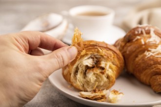 Croissant on white plate with hand on brown concrete background and linen textile, cup of coffee,