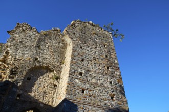 A crumbling stone tower rises under a clear blue sky with few plants, Mystras, Mistra, UNESCO World