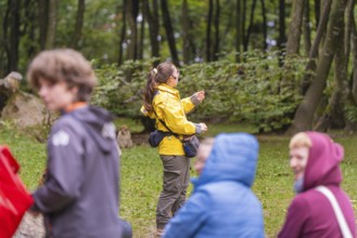 A person in a yellow jacket points to something in a wooded area. Surrounded by blurred figures,