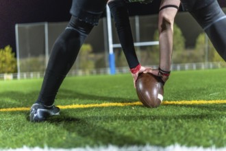 American football player in dark uniform with arm sleeves placing a brown ball on green turf at