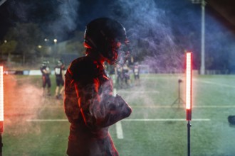 American football player standing on a dark field at night in helmet and pads, framed by red and