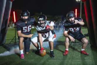 Three determined american football players, one holding the ball, emerging from an inflatable