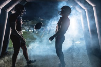 Two american football players wearing helmets and uniforms emerge from a tunnel onto stadium field