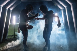 American football players, one holding a ball, emerging from a smoky stadium tunnel at night,