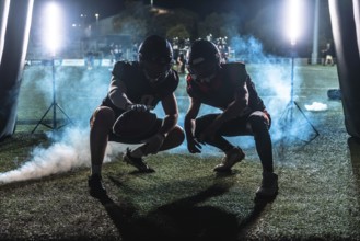 Two american football players in full gear, crouching and ready for action, emerging from a dark