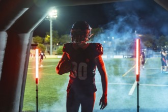 American football player in helmet and uniform bursts from a smoke filled tunnel onto a floodlit